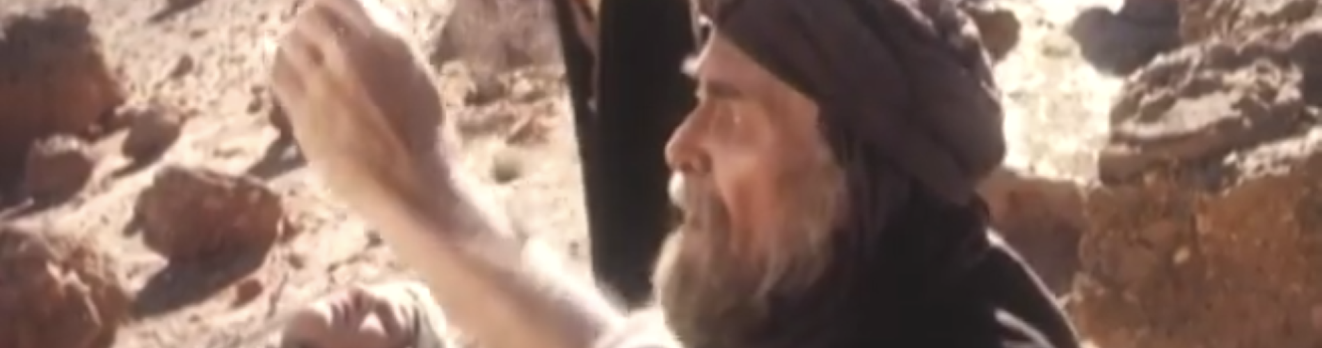 Man in traditional clothing kneeling beside another man lying on rocky ground, raising hand in distress.