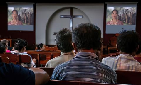 People seated in a church watching a video projected on two screens near a large cross.