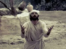 Man in ancient clothing kneels outdoors with arms raised, appearing to plead or pray.